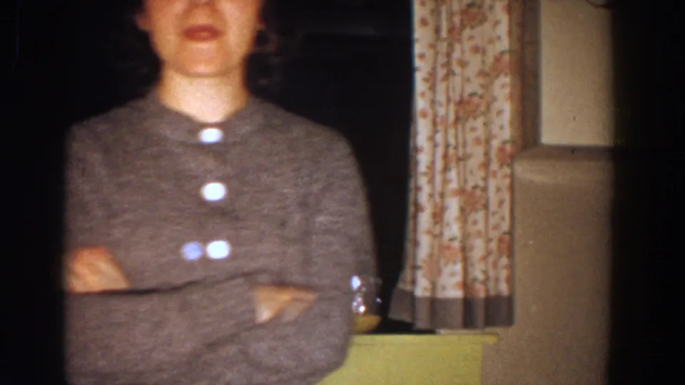 In a 1958 Aliquippa home-movie frame, a woman in a gray buttoned sweater stands indoors with arms crossed beside floral curtains.