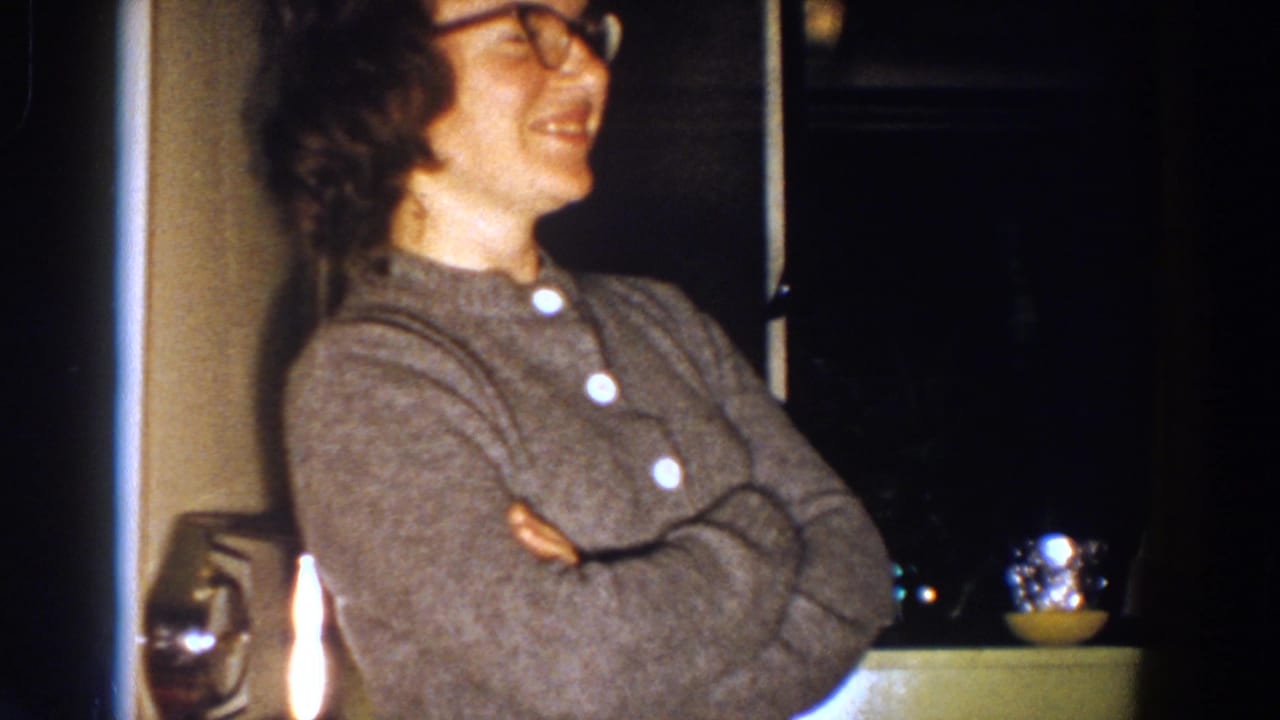 The woman, wearing glasses and a gray sweater, smiles in profile inside a dim Aliquippa domestic room in 1958 footage.