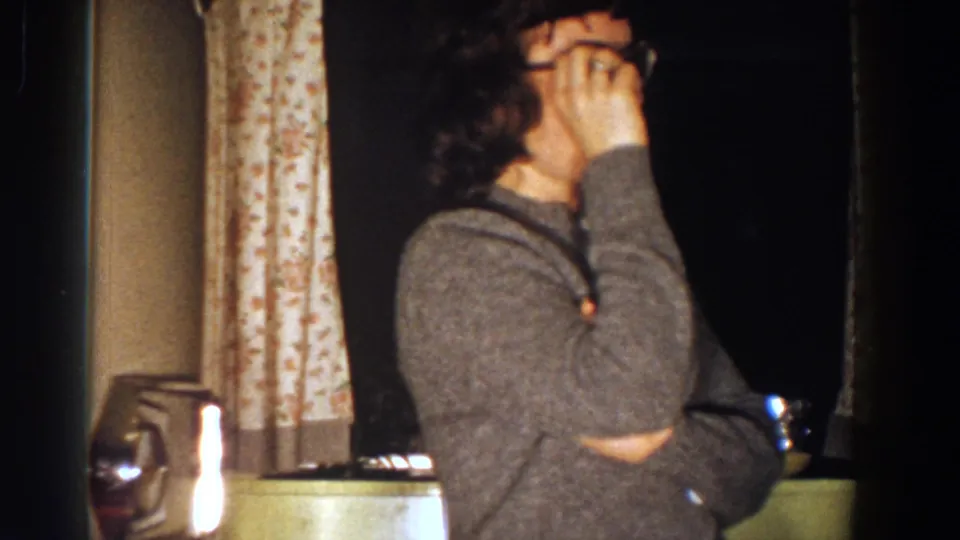 A woman in glasses turns sideways and touches her face while standing near floral curtains in the 1958 Aliquippa home-movie scene.