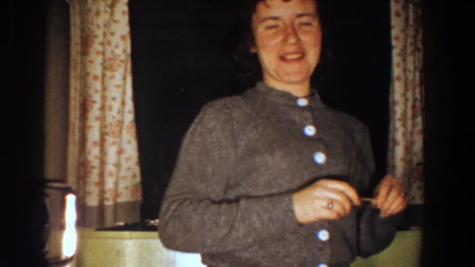 The woman faces the camera in a gray buttoned sweater, appearing to speak or smile in a dim 1958 Aliquippa interior.