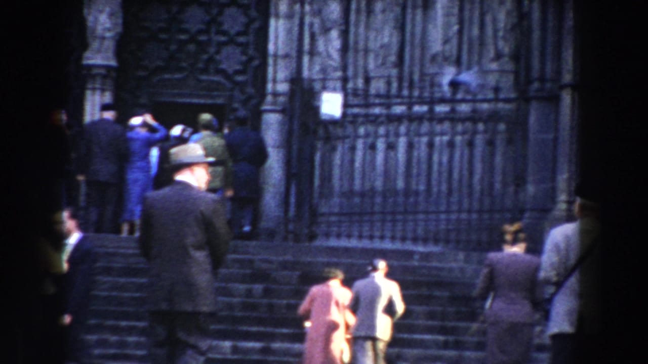 1959 Barcelona home-movie frame of pedestrians ascending stone steps toward an ornate public or religious entrance in damp weather.