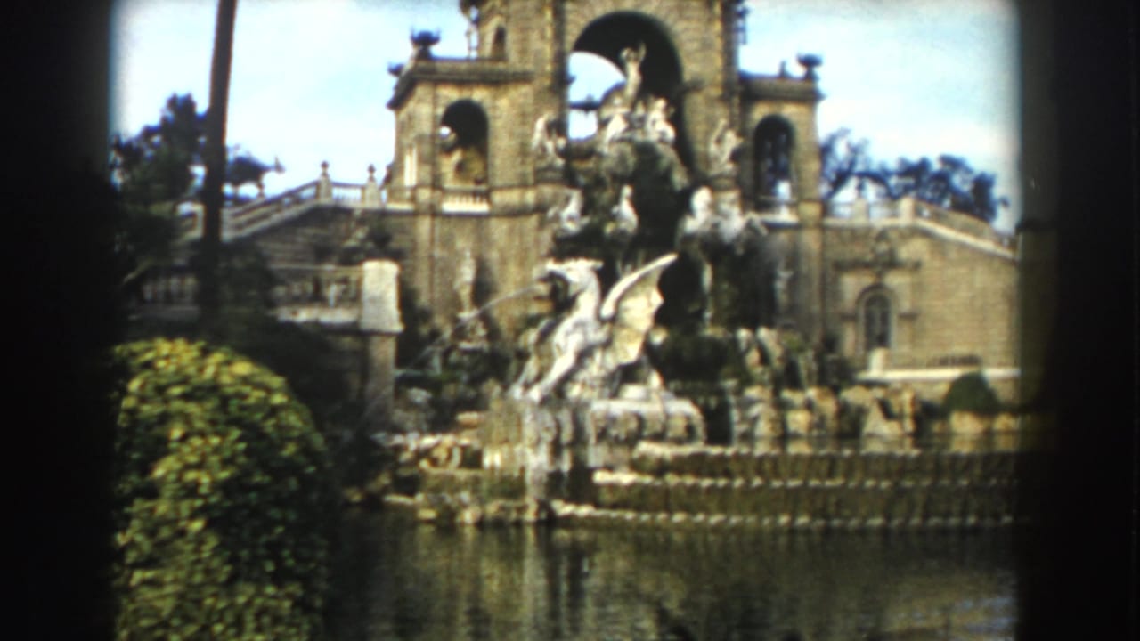 An ornate sculptural fountain stands beside water, hedges, stairs, and arched stone architecture.