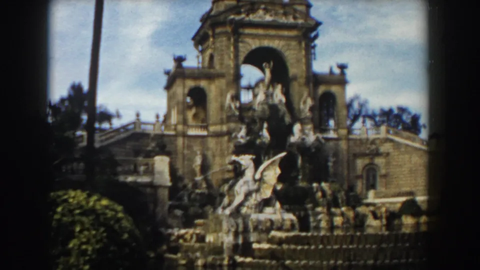 Late-1950s Barcelona 8mm frame of the ornate fountain from a slightly darker angle, with sculpture, water, stairs, and arches visible.