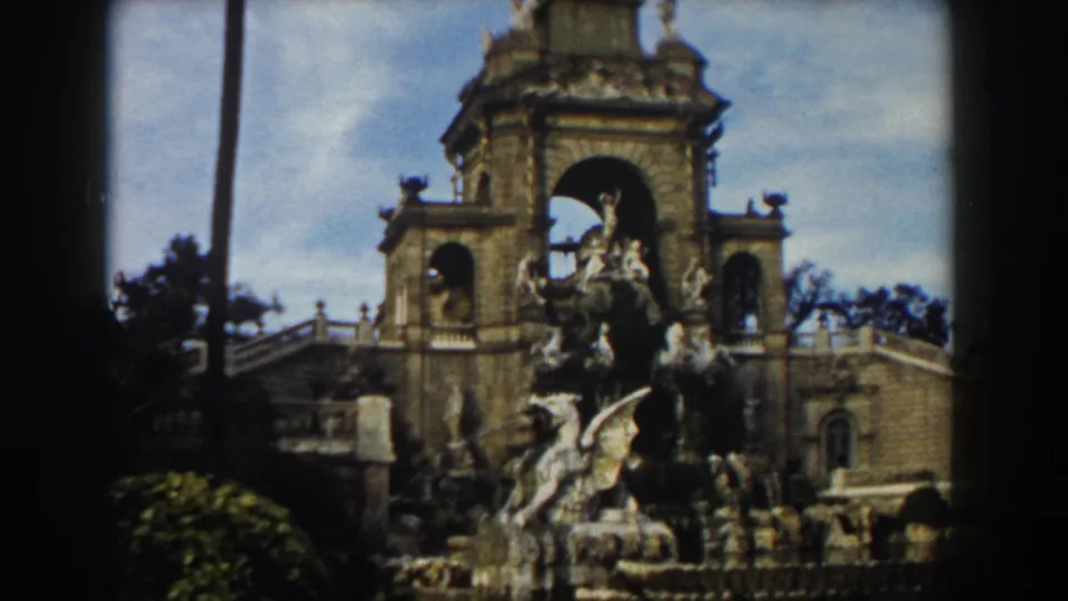 1959 Barcelona home-movie frame showing an ornate stone monument with arches, sculptural figures, palm trees, and faded color.