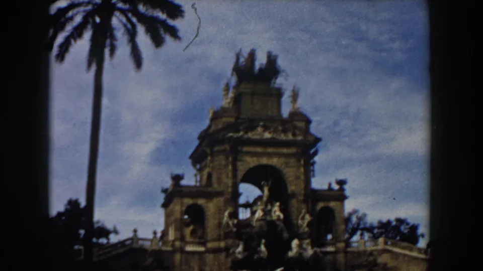 1959 Barcelona home-movie frame showing the monument from a lower angle with a tall palm tree and soft, grainy color.