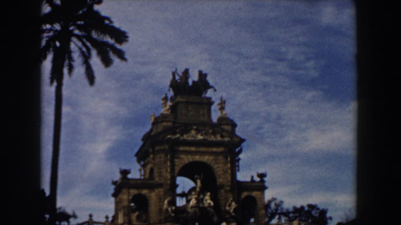 1959 Barcelona home-movie frame showing a palm tree framing an ornate stone monument with arches and sculpted figures.