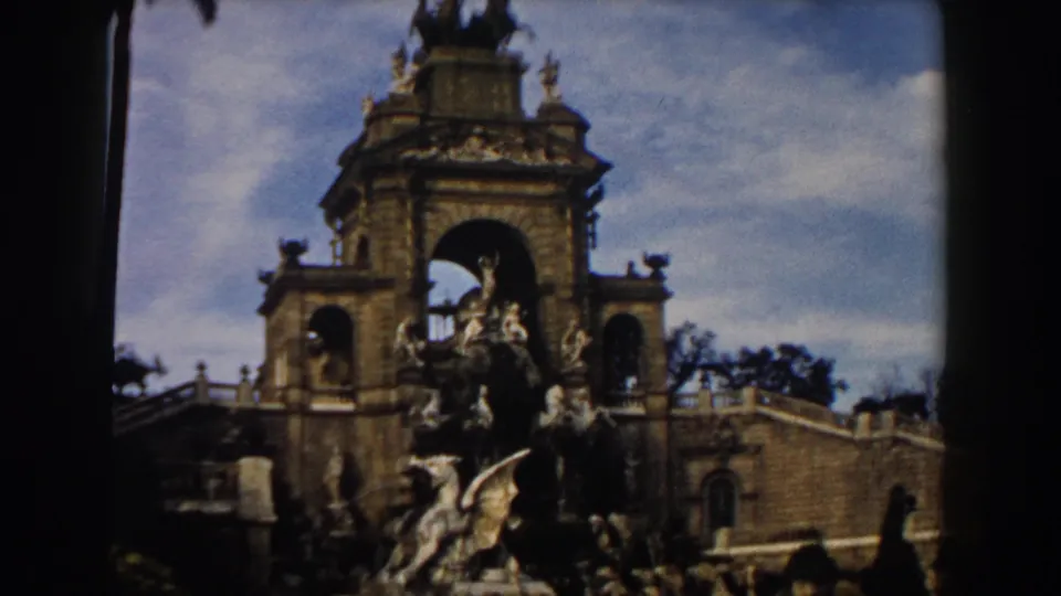 1959 Barcelona home-movie view looking up at the monument, with sculpted figures, stair walls, fencing, and visitors near the base.