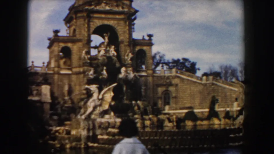 1959 Barcelona frame showing visitors near the monument's base, with carved stone figures, arched openings, fencing, and a curved wall.