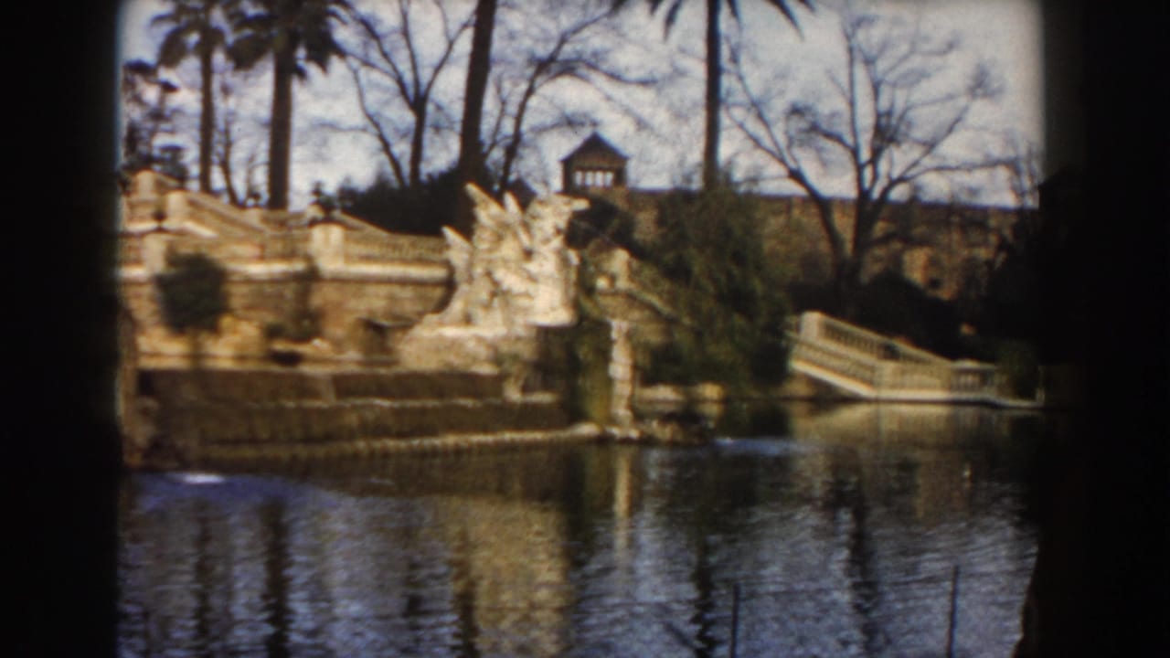 1959 Barcelona home-movie view of an ornate fountain, stone figures, stairs, calm water, palm trees, and bare branches.