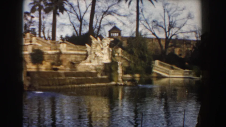 1959 Barcelona home-movie view of an ornate fountain, stone figures, stairs, calm water, palm trees, and bare branches.
