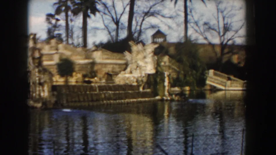 1959 Barcelona 8mm frame showing a stone fountain terrace, sculptural figures, water reflections, palms, and winter-bare trees.