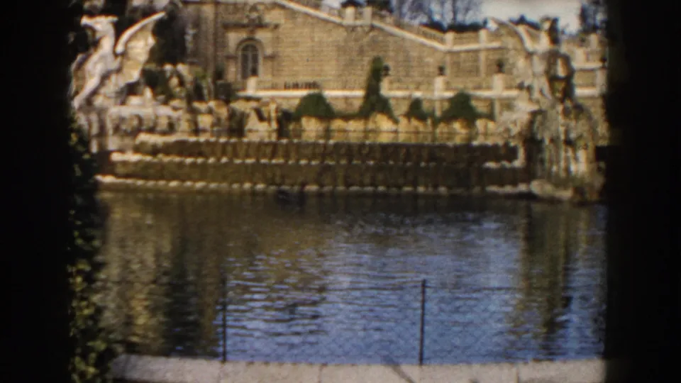1959 Barcelona home-movie frame with stone balustrade, fountain sculptures, small distant visitors, and calm water in front.