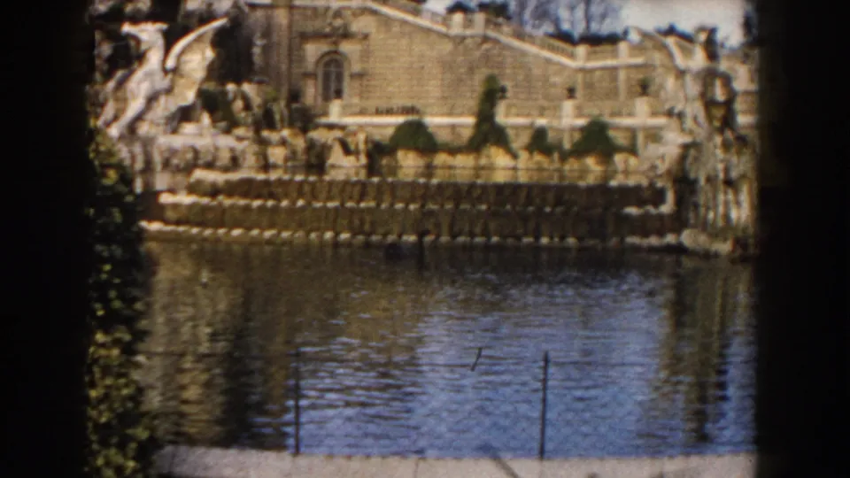 1959 Barcelona 8mm view of the fountain wall, sculptural stonework, small distant visitors, water, and thin fencing at the edge.