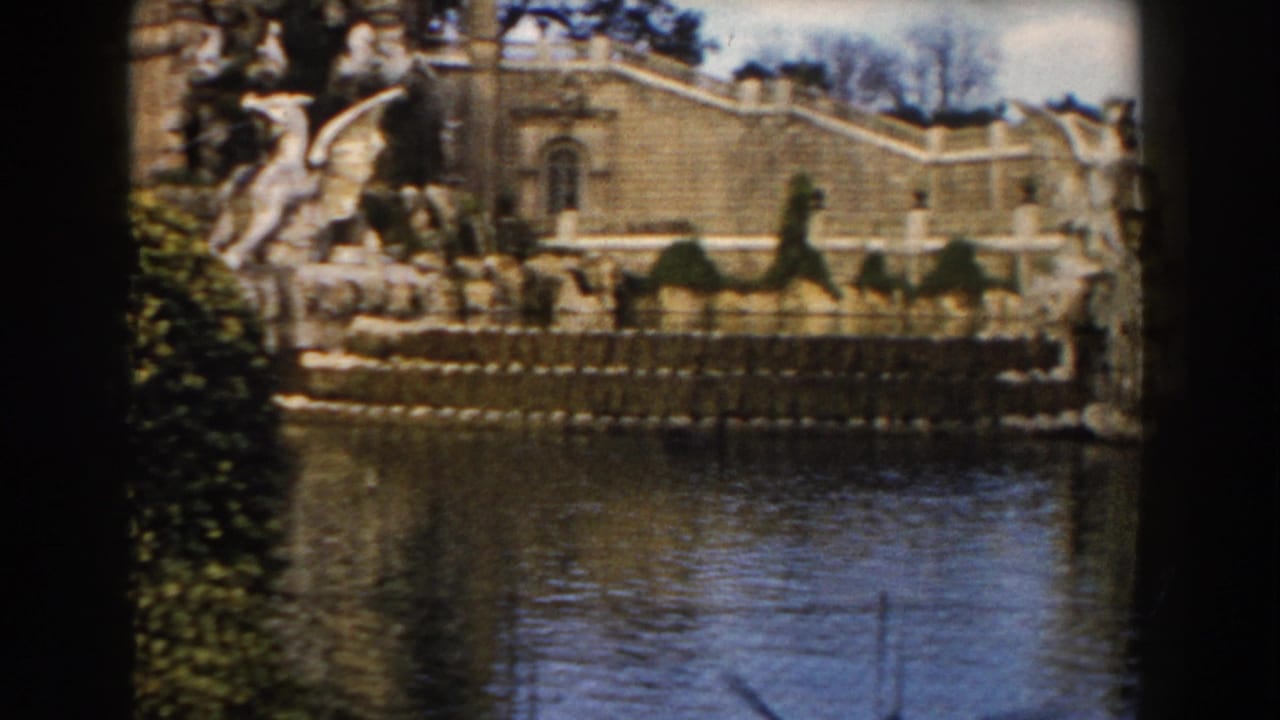 1959 Barcelona home-movie frame showing an ornate stone fountain, formal hedges, and historic architecture across water.