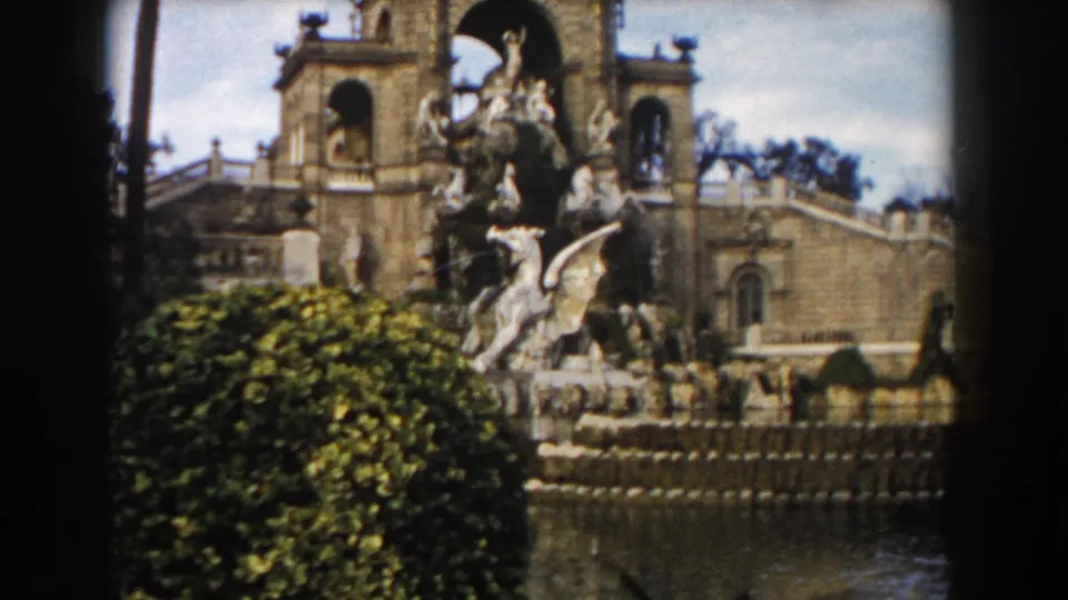 1959 Barcelona home-movie view of ornate fountain statuary framed by greenery, water, and a historic building with arched openings.