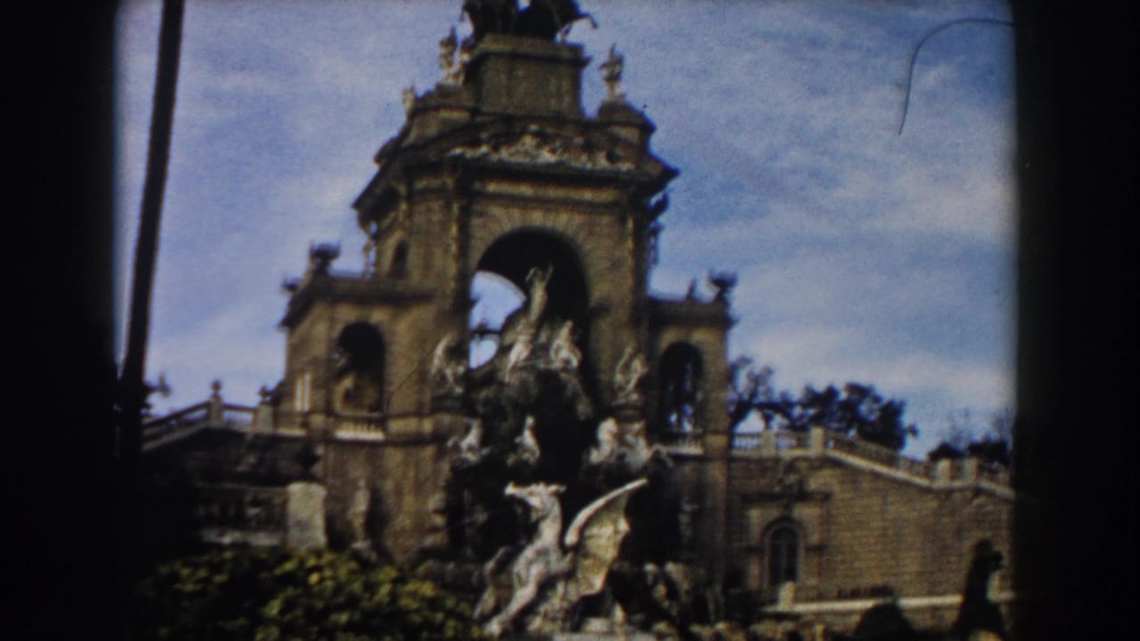 1959 Barcelona 8mm frame showing an ornate stone fountain, arched architectural backdrop, sculpture groups, palm trunks, and blue sky.