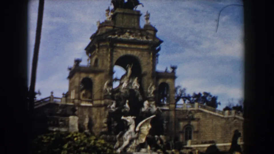 1959 Barcelona 8mm frame showing an ornate stone fountain, arched architectural backdrop, sculpture groups, palm trunks, and blue sky.