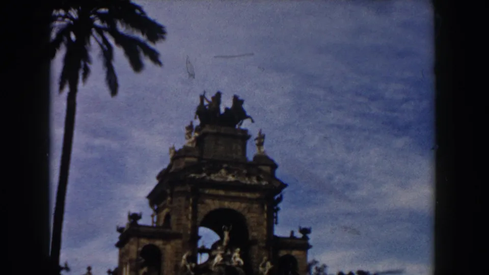 1959 Barcelona 8mm frame showing a palm trunk, blue sky, and the upper portion of an ornate fountain monument with sculptural details.