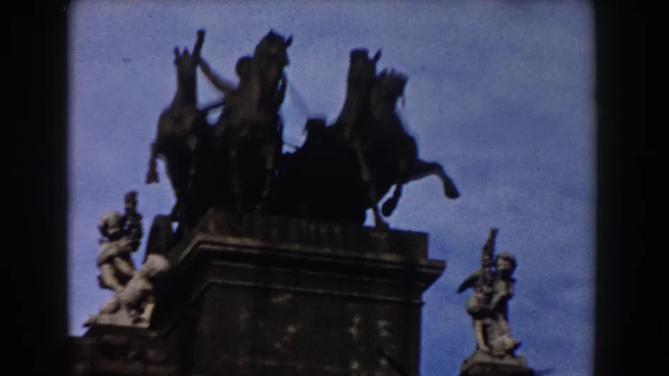 1959 Barcelona frame showing an equestrian statue on a high pedestal, adjacent sculpture figures, and blue sky in soft 8mm focus.