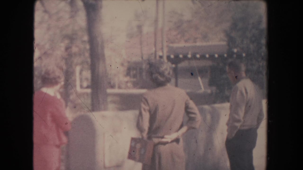 In 1951 New Mexico home-movie footage, three adults stand on a sidewalk near a low wall, facing away toward something offscreen.