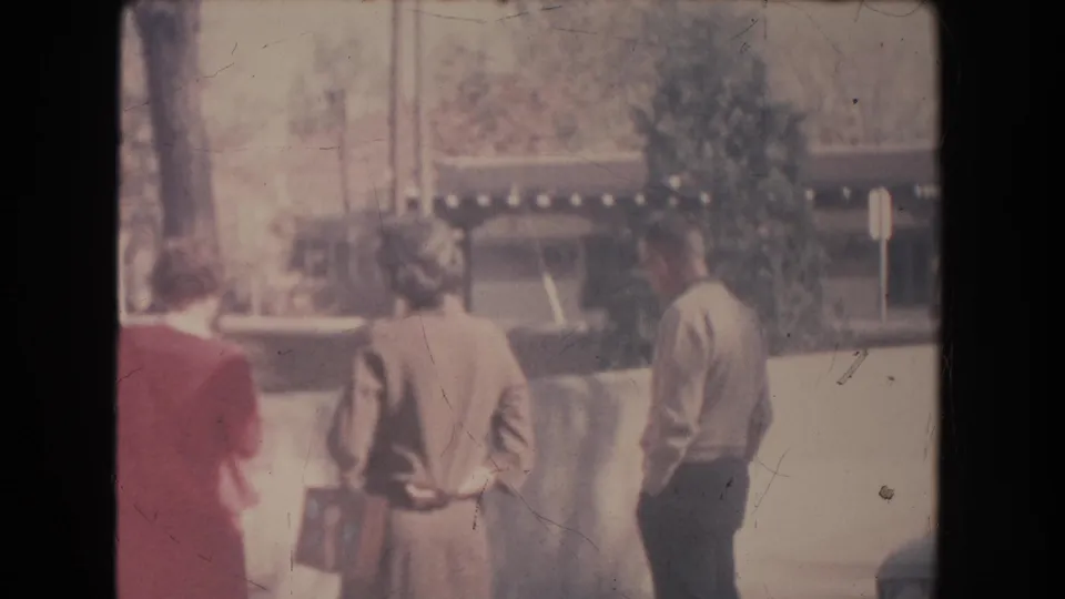 Three adults in mid-century clothing stand near a low wall in New Mexico, with trees, a signpost, and a shaded building behind them.