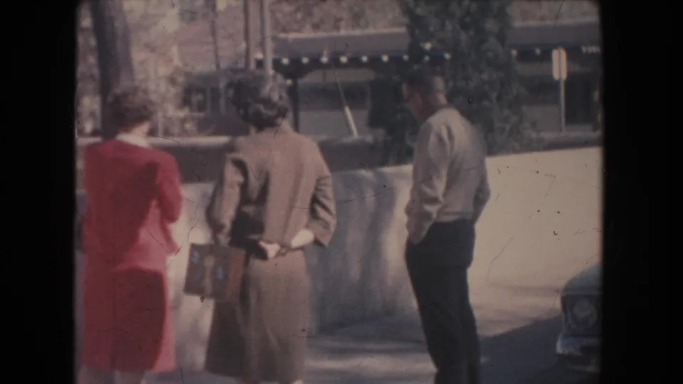 Two women and a man stand along a sidewalk by a low wall, with a parked car edge entering the lower right of the frame.
