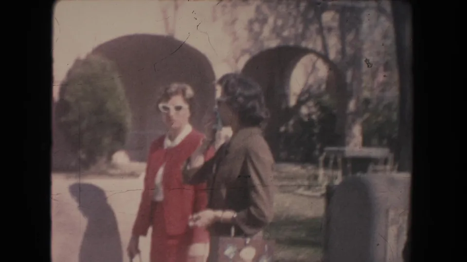 1951 New Mexico frame with two women walking near an arched building, one in a red suit and white sunglasses.