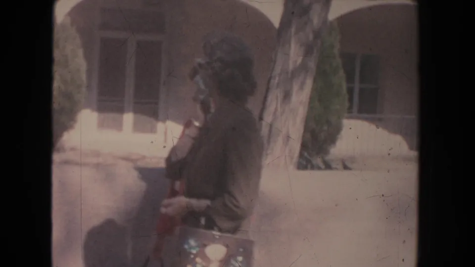 1951 New Mexico frame with a woman in a brown jacket and patterned purse moving past a tree and building facade.