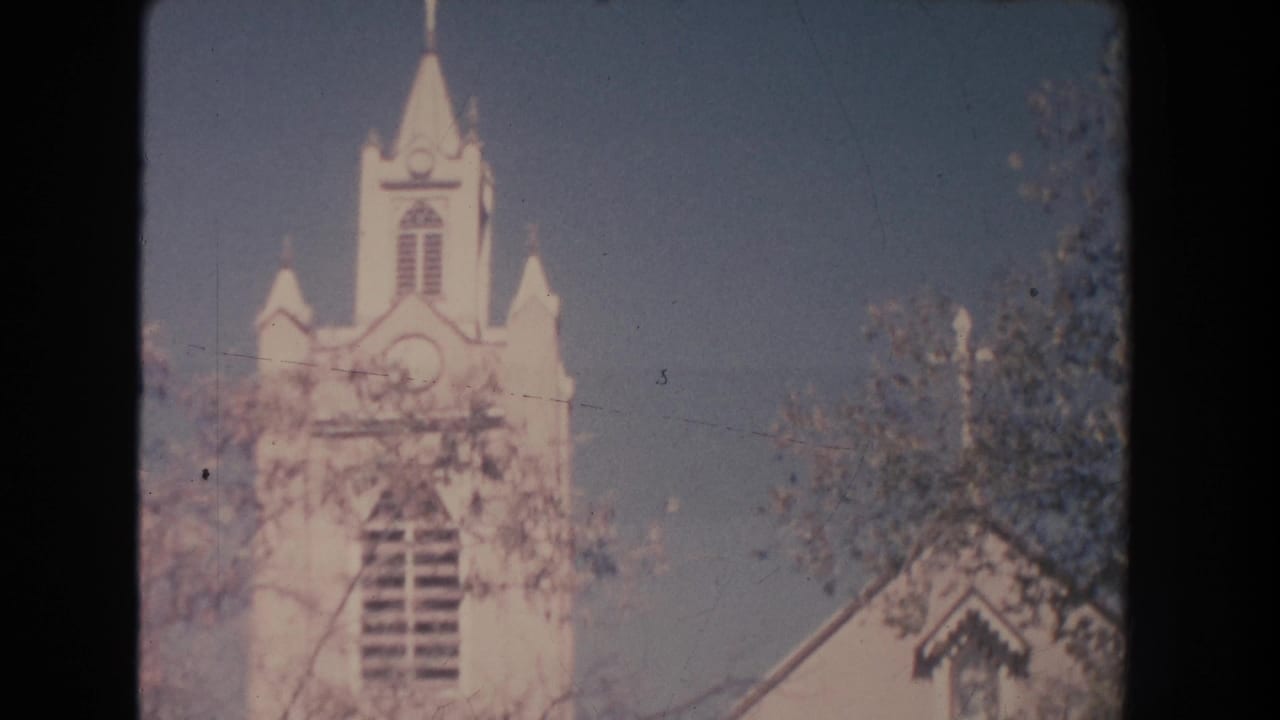 1951 New Mexico 8mm frame showing a white church steeple and facade partly screened by trees in bright daylight.