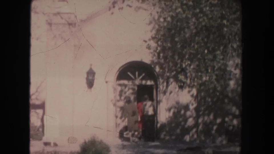 White building entrance with an arched doorway, wall lantern, shrubs, and a person near the door in a 1951 New Mexico village scene.