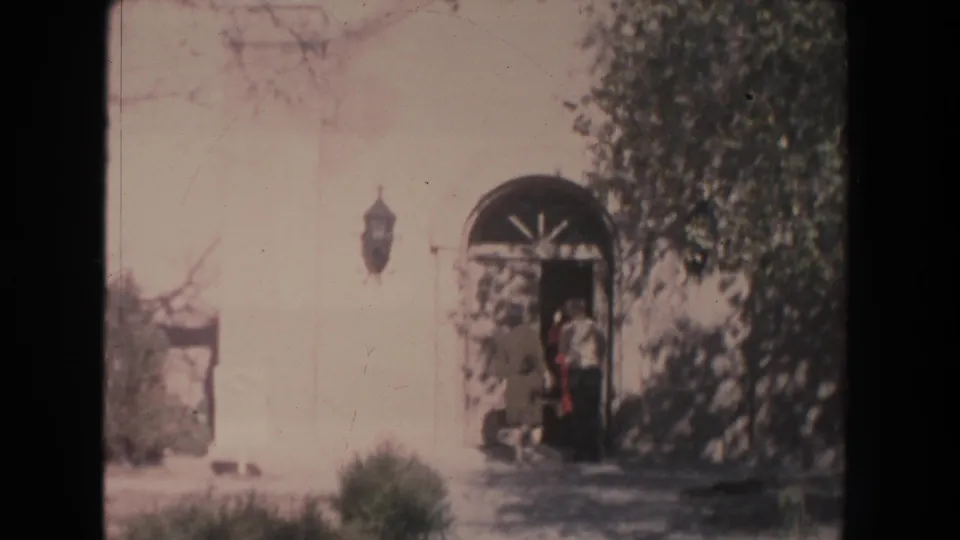 1951 New Mexico home-movie frame of people near an arched doorway on a white building, with shrubs and tree shade around the entrance.