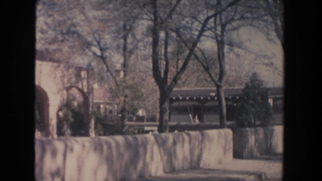 1951 New Mexico 8mm frame showing a low stone wall, bare trees, shrubs, and a white building with arched openings.