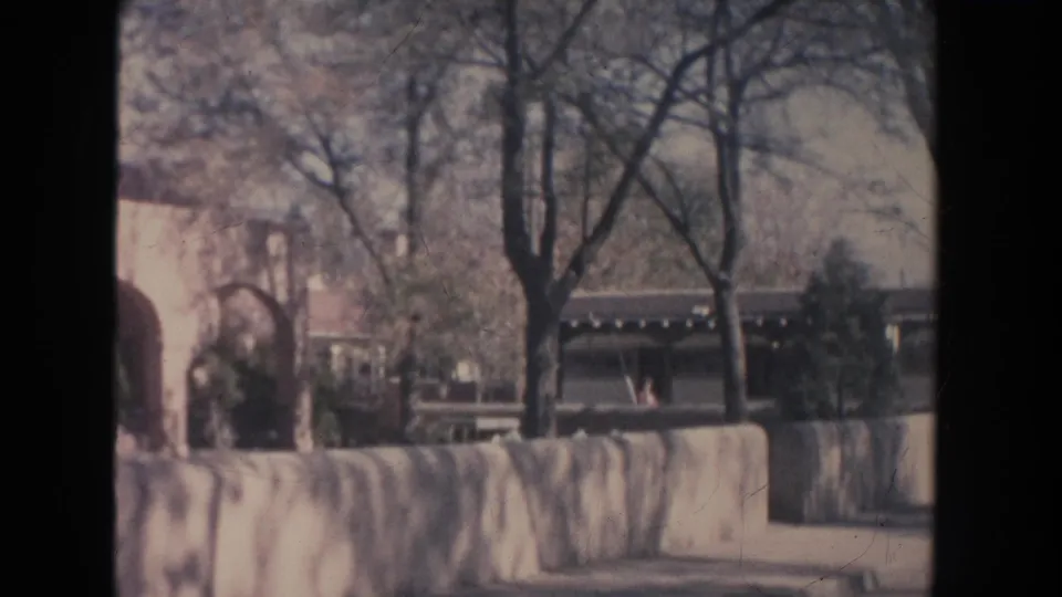 1951 New Mexico 8mm frame showing a low stone wall, bare trees, shrubs, and a white building with arched openings.
