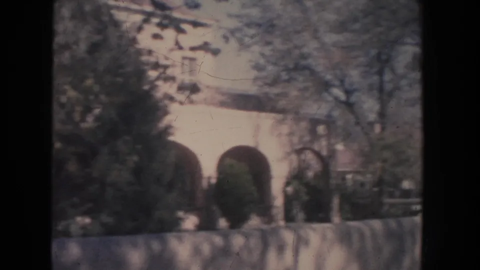1951 New Mexico home-movie frame with arched white architecture partly obscured by branches, shrubs, and a low wall.