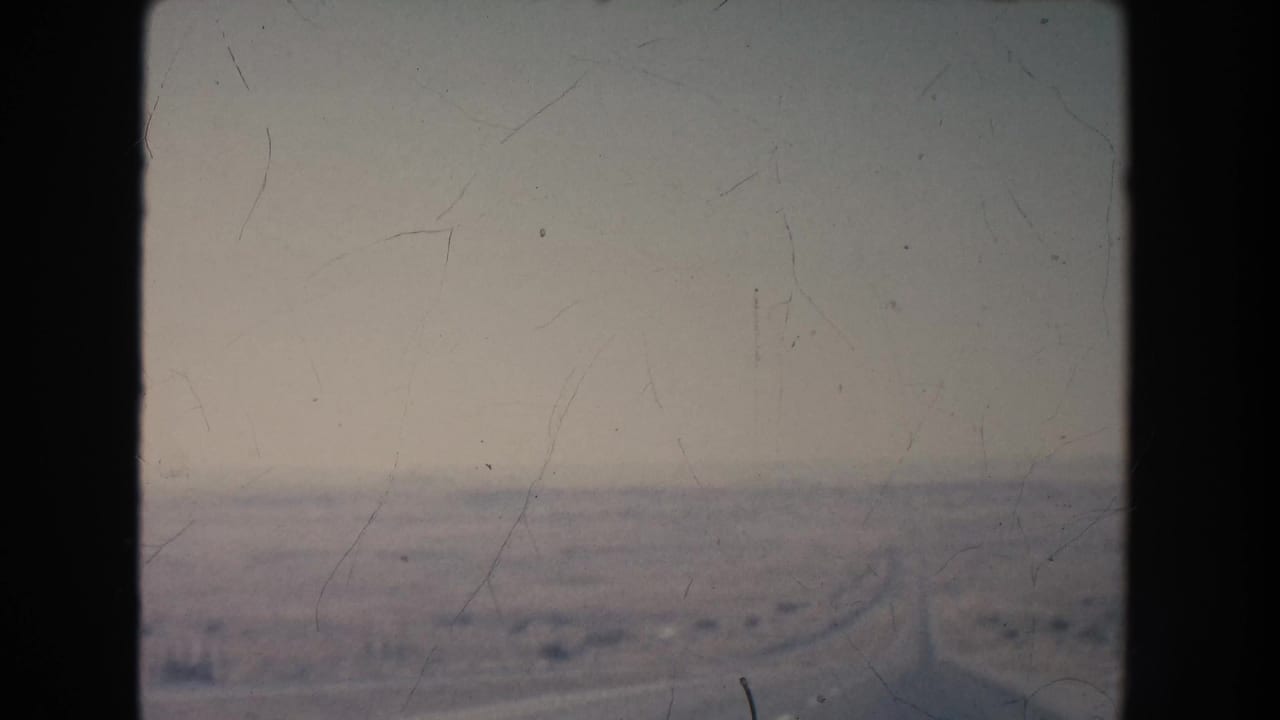 1951 8mm view of a long road crossing a hazy New Mexico desert plain with sparse vegetation and low hills in the distance.