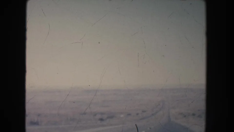 1951 8mm view of a long road crossing a hazy New Mexico desert plain with sparse vegetation and low hills in the distance.