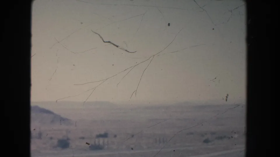 Soft 8mm frame of New Mexico desert terrain, with low hills on the horizon and scattered scrub near the road edge.