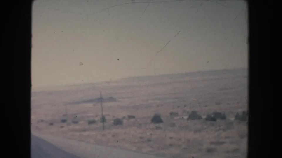 Soft 8mm frame of a New Mexico desert road edge, with a utility pole, scattered scrub, and low hills under a washed pale sky.