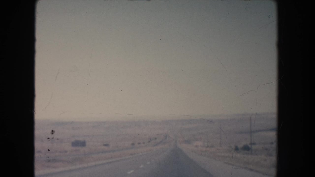 1951 New Mexico 8mm frame showing a road-ahead view across arid terrain toward faint mesa formations and roadside poles.