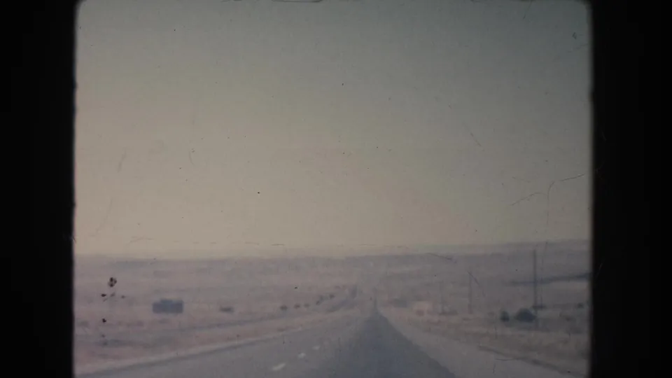 1951 New Mexico 8mm frame showing a road-ahead view across arid terrain toward faint mesa formations and roadside poles.