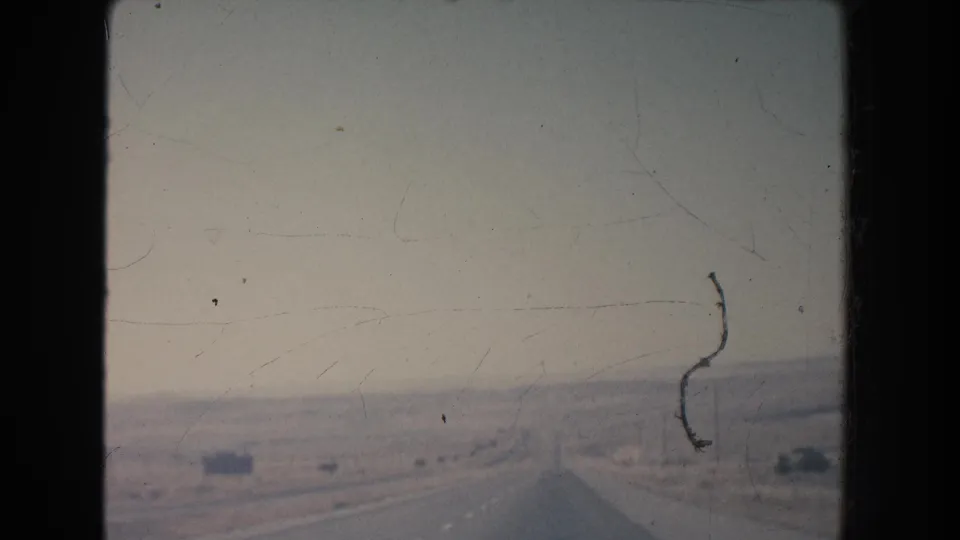 Forward-moving 1951 New Mexico road view with a flat desert shoulder, faint mesa shapes, roadside poles, and visible 8mm film wear.