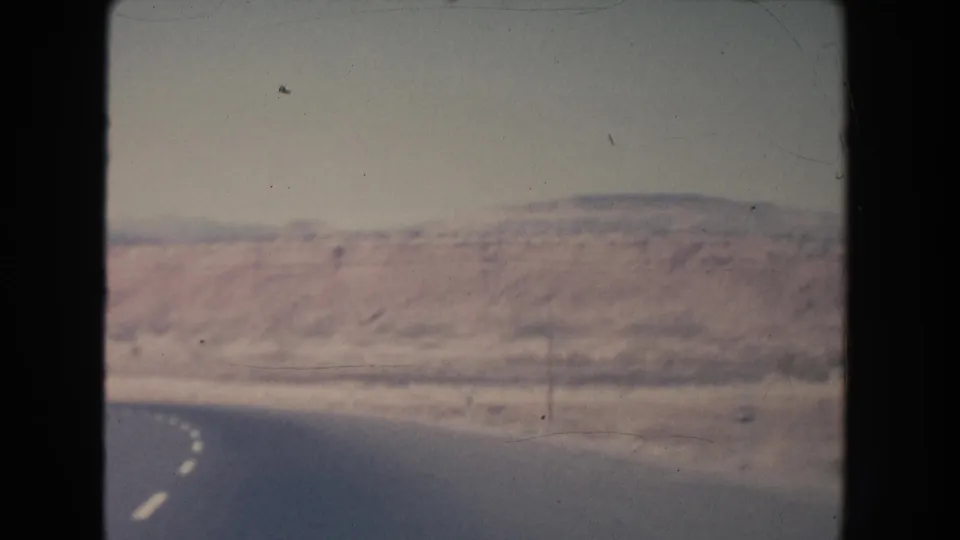 1951 New Mexico 8mm frame with the highway curving near a low red mesa-like rock formation and sparse roadside poles.