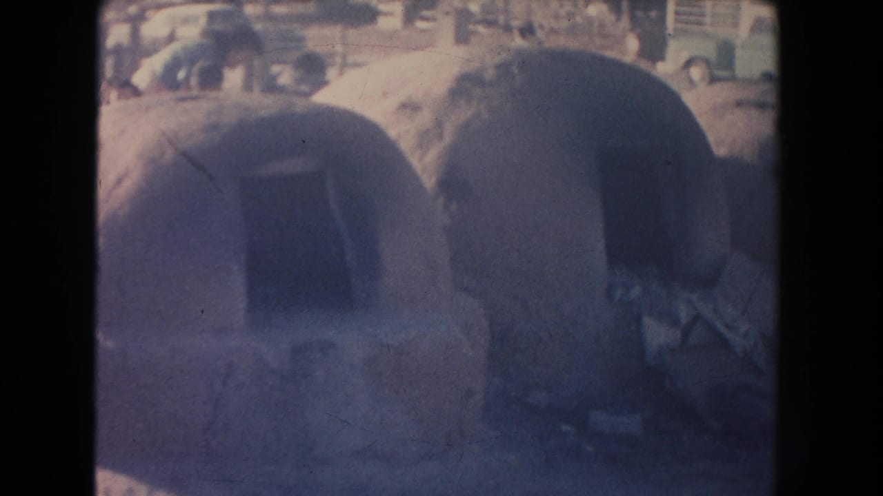 1951 New Mexico home-movie frame showing rounded adobe dwellings with shadowed doorways and faint classic cars in the background.