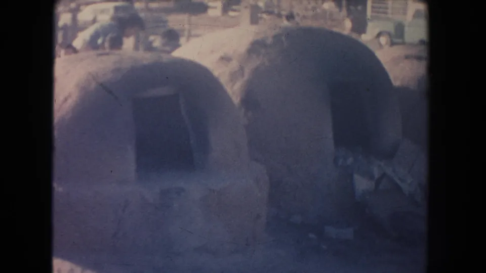 1951 New Mexico home-movie frame showing rounded adobe dwellings with shadowed doorways and faint classic cars in the background.