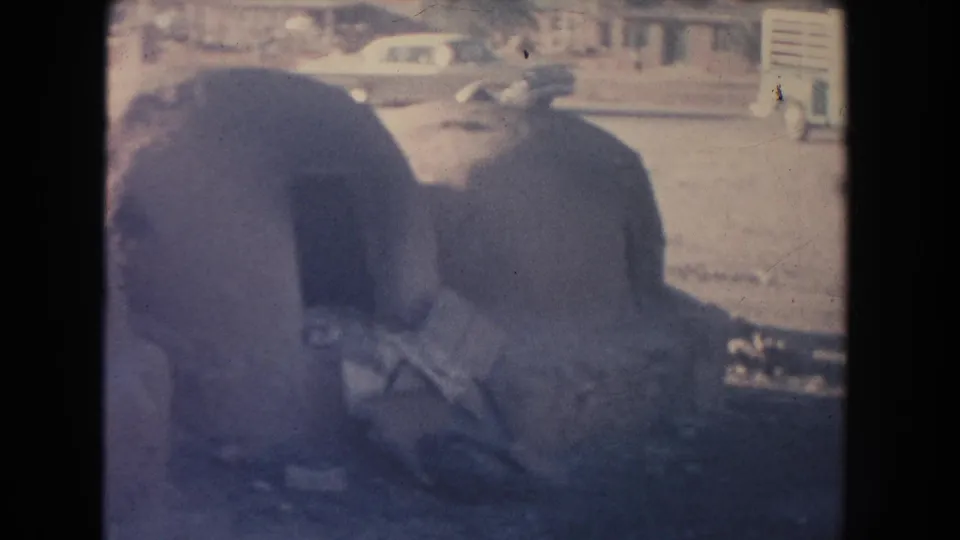 1951 New Mexico roadside view with adobe structures in the foreground, classic cars behind, and a faintly visible person near the road.