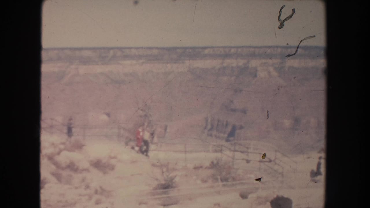 Distant visitors walk along a pale overlook path with railings and canyon terrain behind them.