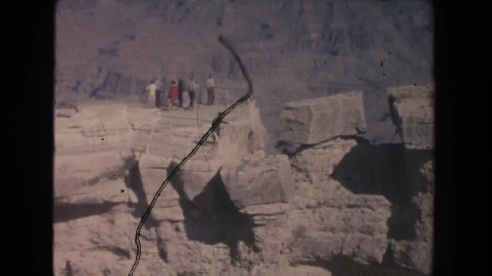 1951 New Mexico home-movie frame showing distant visitors at a canyon edge with pale rock walls and a dark chasm below.