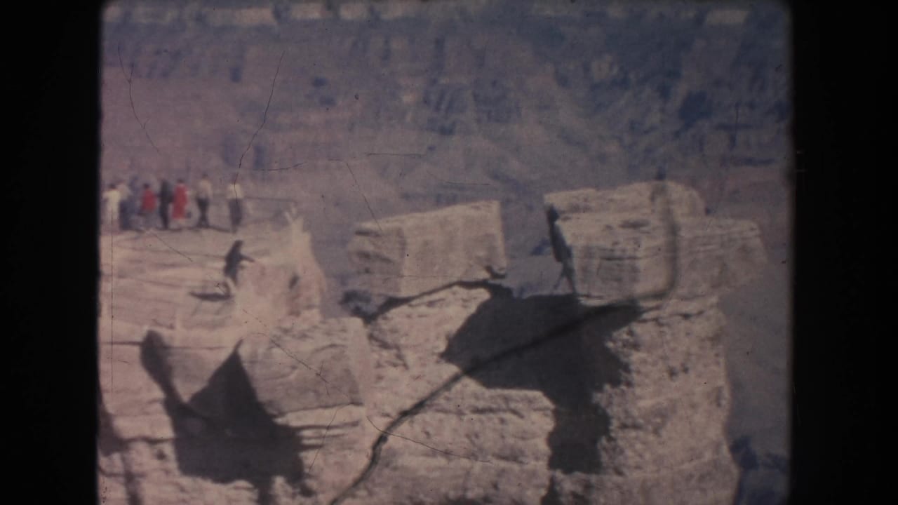 Small figures stand near a canyon edge with layered rock beyond.