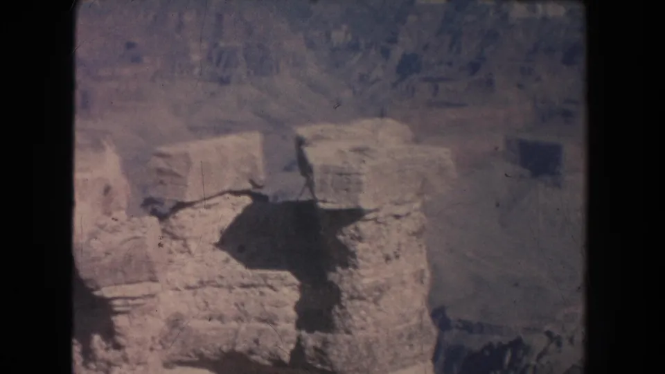 Faded 8mm frame of a New Mexico canyon overlook, emphasizing pale stone, a dark chasm, and distant visitors at the edge of the frame.