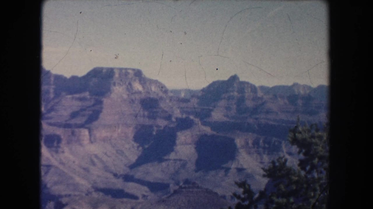 Wide canyon landscape with layered cliffs, pale sky, and tree edge.